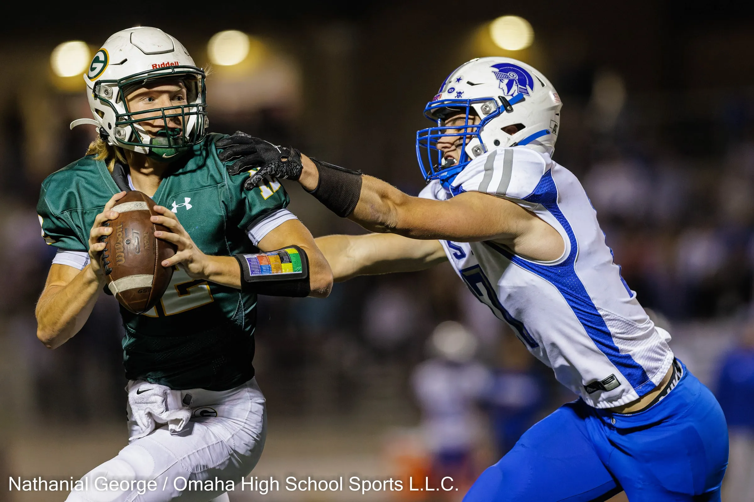 A quarterback in green with a G on his helmet holds the ball and looks downfield as a defender in white and blue reaches an arm across in front of him, under stadium lights.