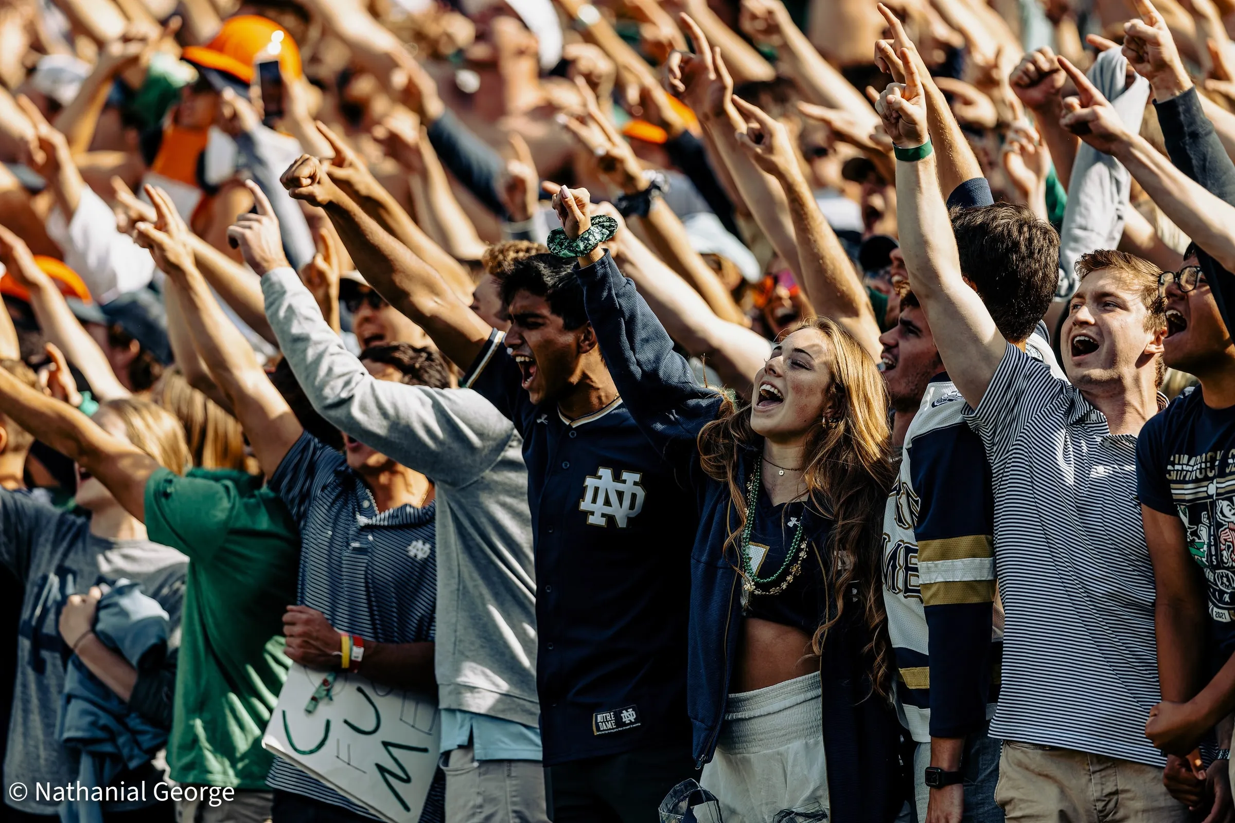 Notre Dame fans pack the stands in bright sun, fingers pointed skyward, shouting, one of them in a navy jersey with the interlocking ND.