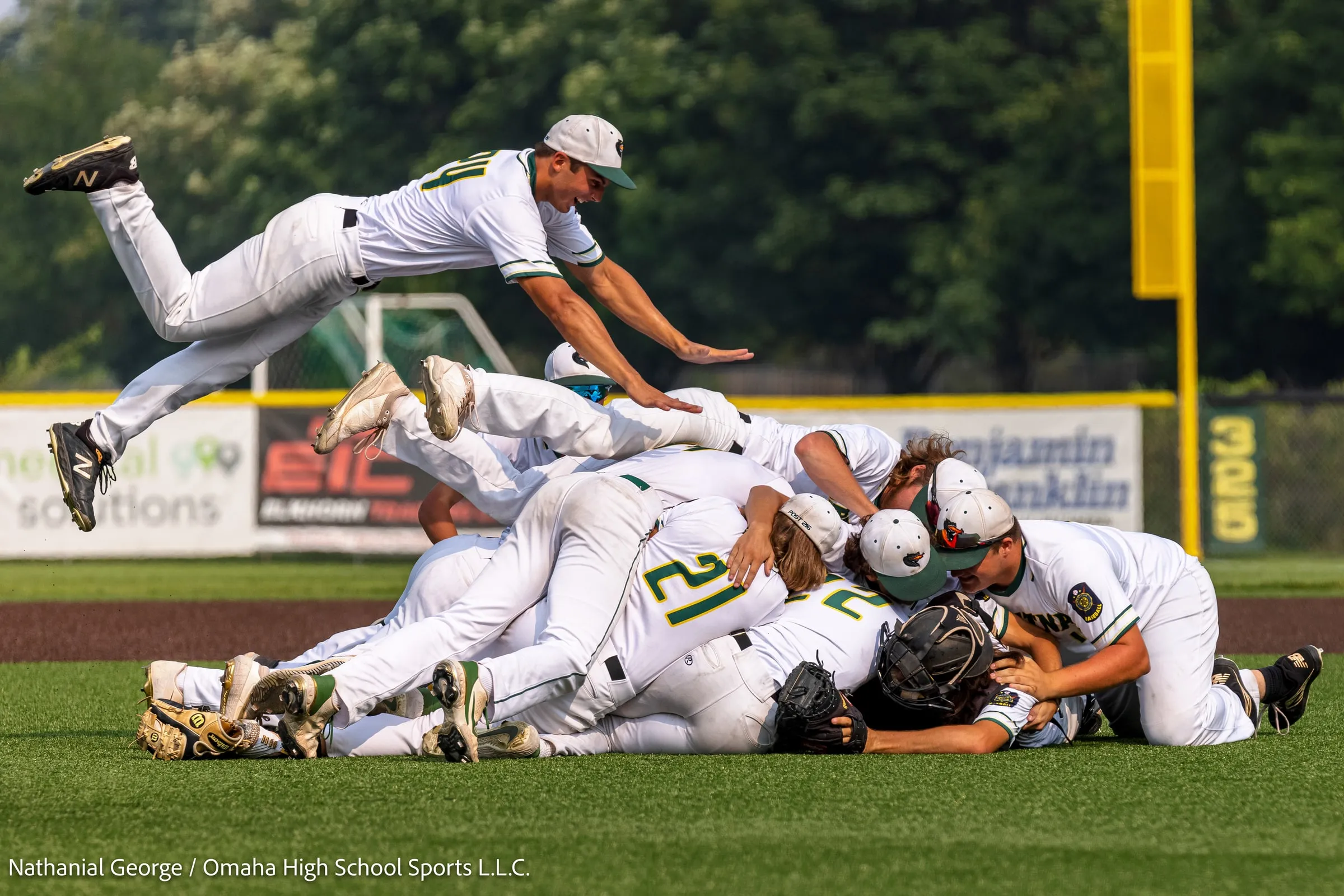 Baseball players in white and green pile onto one another on the infield as a teammate dives over the top, the yellow foul pole and the 325 sign on the fence behind them.