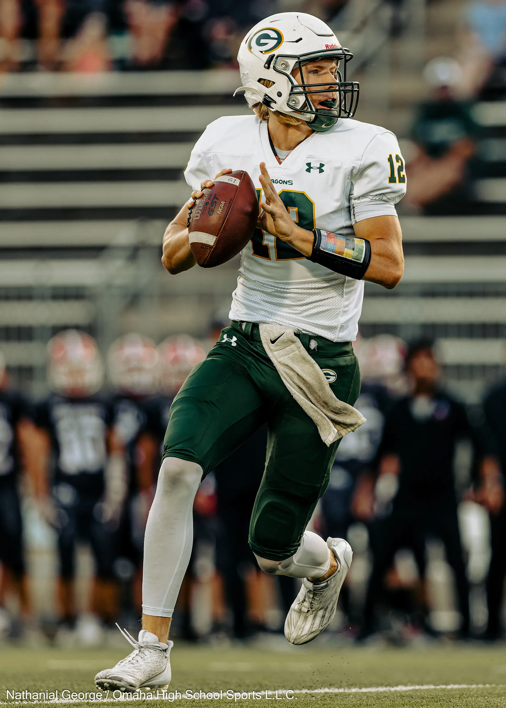 A quarterback in a white Dragons jersey, number 12, and green pants drops back holding the ball in both hands, the opposing sideline in black behind him.