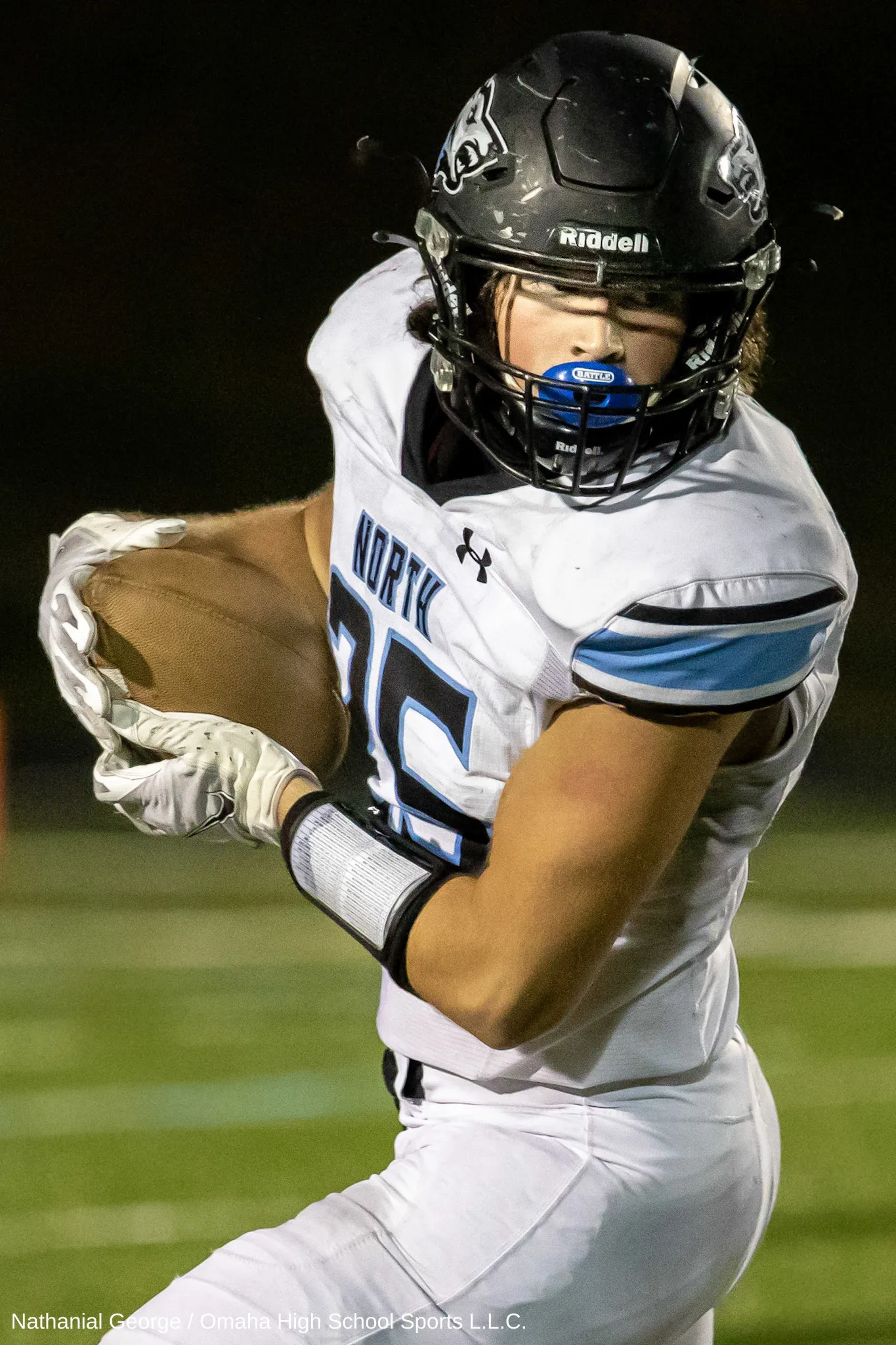 A ball carrier in a white North jersey and a black helmet tucks the ball under his arm at night, a blue mouthguard clenched in his teeth.