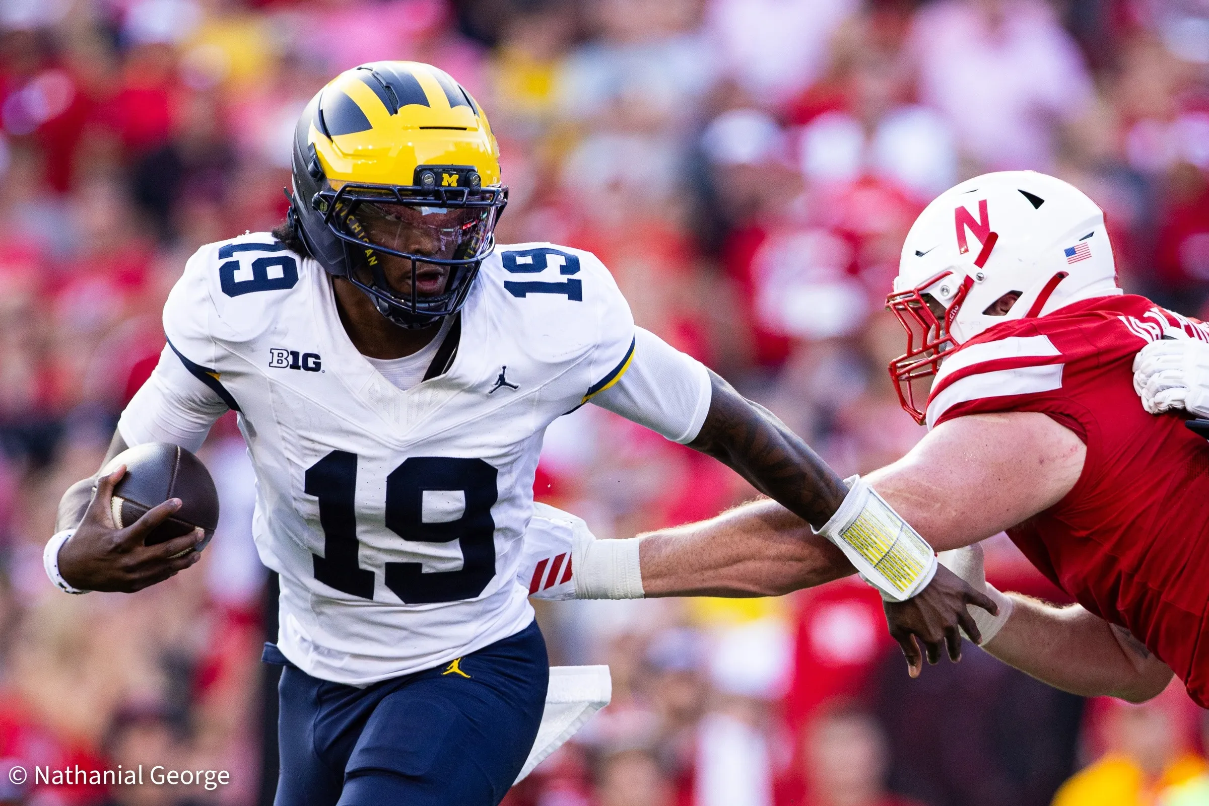 A quarterback in white, number 19, runs with the ball tucked as a defender in red reaches across his arm from the right.