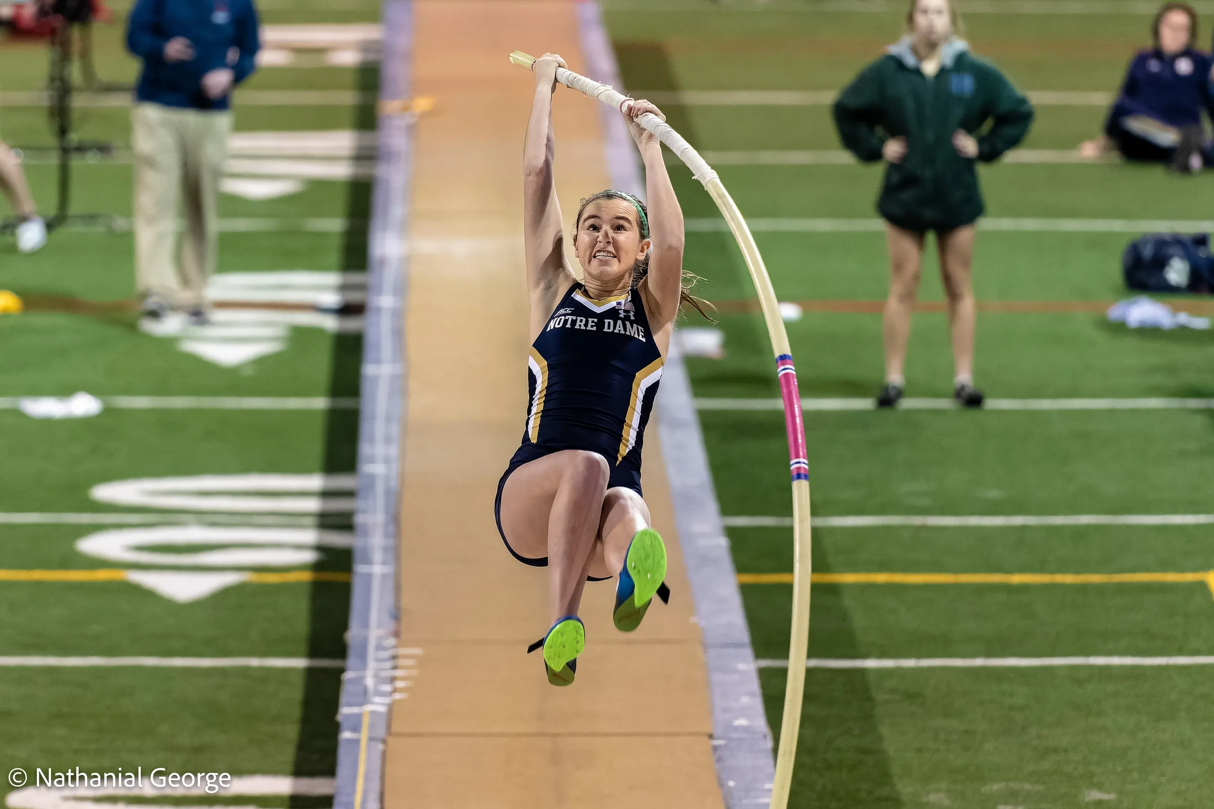 A Notre Dame pole vaulter in navy leaves the runway hanging from the bending pole, knees tucked and green spikes in the air, as others watch from the infield.