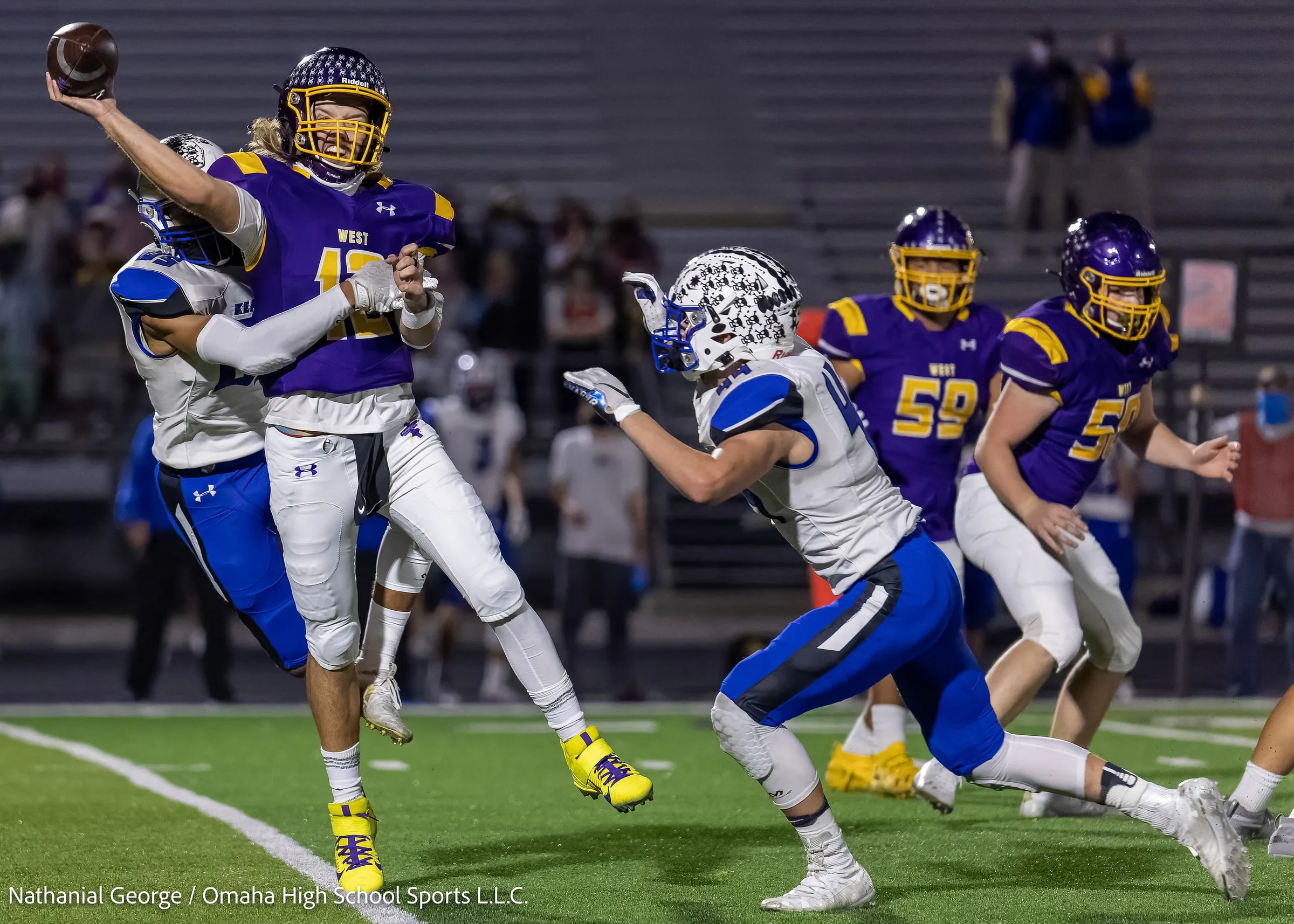 A quarterback in purple and gold gets a pass away as a defender in white and blue hits him from behind and a second defender rushes in with his arms out, under the lights.